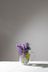 Beautiful small meadow bouquet in a transparent mug on a white-gray background with copy space. Minimalism.