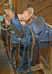 Two retro leather horse saddles, one brown and the other a light tan color, on sawhorses with various iron brands in the background.
