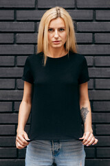 Stylish blonde girl wearing black t-shirt and glasses posing on black wall background, urban...