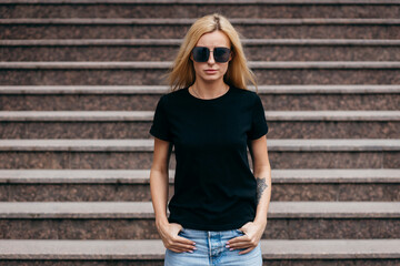 Stylish blonde girl wearing black t-shirt and glasses posing against street , urban clothing style....