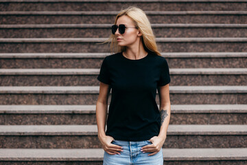 Stylish blonde girl wearing black t-shirt and glasses posing against street , urban clothing style. Street photography - obrazy, fototapety, plakaty