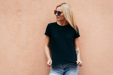 Stylish blonde girl wearing black t-shirt and glasses posing against street , urban clothing style. Street photography - obrazy, fototapety, plakaty