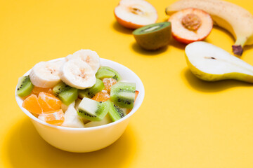 fruit salad in a small white bowl on a yellow background, cut fruit in the background in blur