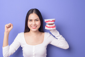 Young beautiful smiling woman is holding plastic denture teeth over isolated purple background studio