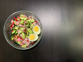 In the photo, a salad of chicken egg, radish, cucumber on a dark wooden table. Photo from above. Proper nutrition, vegetarian diet.