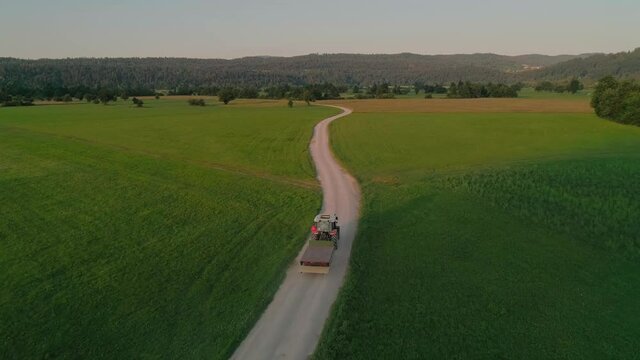 Cinematic aerial drone shot of camera follow small agricultural tractor on gravel country road. Beautiful and peaceful image of country farm life at sunset. Fertile soil and earth for sustainable farm