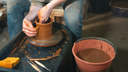 Fototapeta na wymiar Man holding crafting tool and working. Craftsman at kick wheel in pottery. Working process of clay item in workshop.