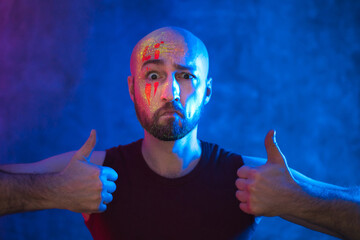 Bald man with makeup shows thumbs up. Fluorescent paint in neon light.