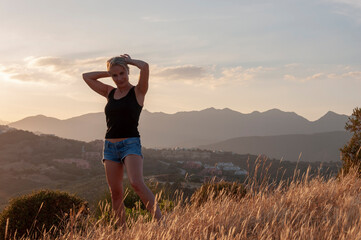 A blonde girl is standing with her hands hair in the grass on top of a hill on a background of mountains during sunset