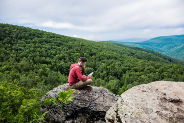 Young man in a red sweater and beige pants sits on top of a high cliff and reads an e-book. Green forest is spread down below. Low mountains overgrown with tall trees can be seen on the horizon.