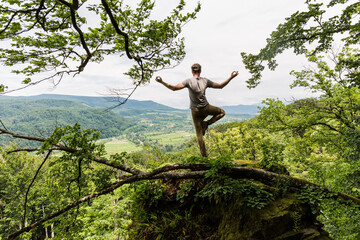 Young man in a grey T-shirt and beige pants stands on one leg in a meditation pose on a high rock...