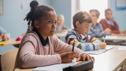 In Elementary School Classroom Brilliant Black Girl Writes in Exercise Notebook, Taking Test and Writing Exam. Junior Classroom with Diverse Group of Children Working Diligently and Learning New Stuff - obrazy, fototapety, plakaty