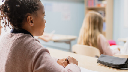 In Elementary School Class: Over the Shoulder View of a Brilliant Black Girl Listening To a Teacher. Junior Classroom with Diverse Group of Bright Children Working and Learning New Stuff