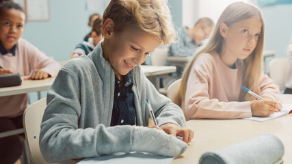 Obraz premium In Elementary School Classroom Brilliant Caucasian Boy Writes in Exercise Notebook, His Hand in a Cast. Junior Classroom with Diverse Group of Bright Children Working Diligently and Learning New Stuff
