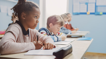 In Elementary School Classroom Brilliant Black Girl Writes in Exercise Notebook, Taking Test. Junior Classroom with Diverse Group of Bright Children Working Diligently and Learning. Side View Portrait