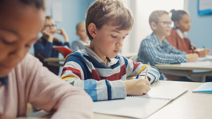 In Elementary School Class: Portrait of Brilliant Caucasian Boy Writes in Exercise Notebook, Taking...