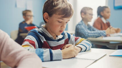 In Elementary School Class: Portrait of Brilliant Caucasian Boy Writes in Exercise Notebook, Taking Test and Writing Exam. Diverse Group of Bright Children Working Diligently and Learning - obrazy, fototapety, plakaty