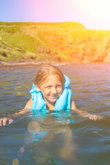 Child in a life jacket on a bright summer day bathes in the sea. Little girl splashing in the water.