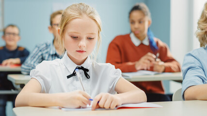 Fototapeta premium In Elementary School Class: Portrait of a Brilliant and Cute Caucasian Girl Writes in Exercise Notebook. Junior Classroom with Diverse Group of Children Working Diligently and Learning New Stuff