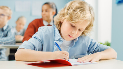 Fototapeta na wymiar In Elementary School Class: Portrait of a Brilliant and Cute Caucasian Boy Writing in Exercise Notebook. Junior Classroom with Diverse Group of Bright Children Working Diligently, Learning New Stuff