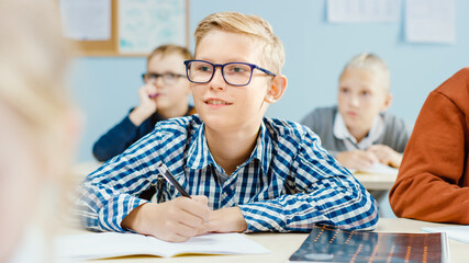 In Elementary School Class: Portrait of a Brilliant Caucasian Boy Wearing Glasses Writes in the Exercise Notebook. Diverse Group of Bright Children Learning Science and Creative Thinking