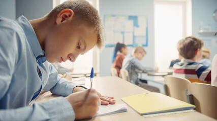 Fototapeta premium Shot of a Young Boy in School Writing in Exercise Notebooks, Taking a Test. Elementary Classroom of Diverse Bright Children Writing in Notebooks.