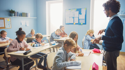 In Elementary School Class: Enthusiastic Teacher walks between Rows of Bright Diverse Children, Dictates Lesson. Group of Smart Multiethnic Kids Writing in Exercise Notebooks. - obrazy, fototapety, plakaty