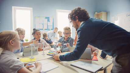 Elementary School Classroom: Enthusiastic Teacher Holding Tablet Computer Explains to a Brilliant Young Children How Wind Turbines Work. Kids Learning about Eco-Friendly Forms of Renewable Energy