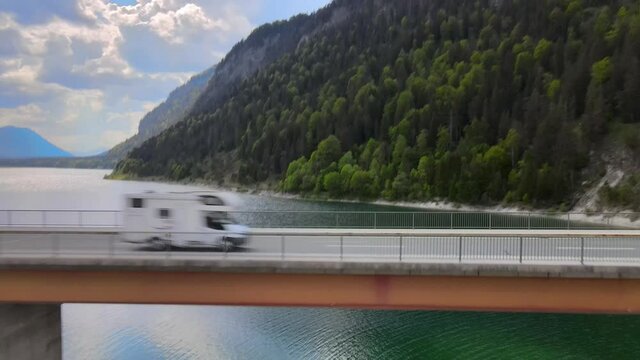 Aerial view flying towards a camper van crossing a bridge over a lake in the alps