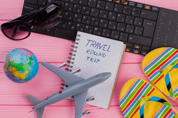 Travel round trip on notebook with woman's traveler accessories glasses wallet and flip-flops on pink table top background. Globe black keyboard grey airplane.