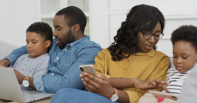 African American parents with kids on couch using gadgets. Young man and woman with son and daughter spending time together with devices Father and son with laptop Mother and daughter with smartphones