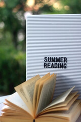 Bulletin board with 'summer reading' caption and open book in a garden. Selective focus.