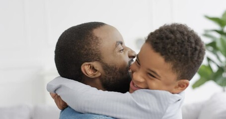 Happy cheerful African American man hugging his small cute son and laughing at home on couch. Joyful parent with little kid in living room. Young father smiling and embracing child indoor. Generations