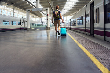 male traveler with a backpack and a suitcase going along the train wagons at railway station.