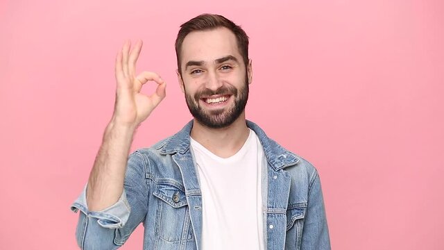 Handsome bearded young man guy 20s years old in denim jacket white t-shirt isolated on pastel pink background studio. People lifestyle concept. Looking camera charming smile showing ok okay gesture
