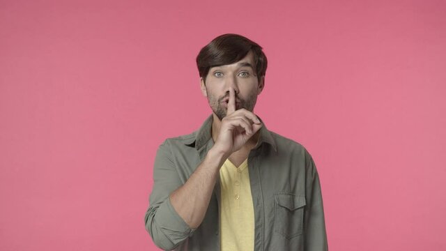 Handsome bearded man asking keep promise and secret safe, shushing with finger pressed to lips, make hush shh sign to tell be quiet or silent, dont speak its taboo, pink background