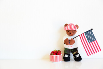 One teddy bear in a red striped hat is standing and holds the flag of America. Light background, indoors. USA Independence Day Background. Picnic on July 4th. Copy space for text. San Francisco, USA