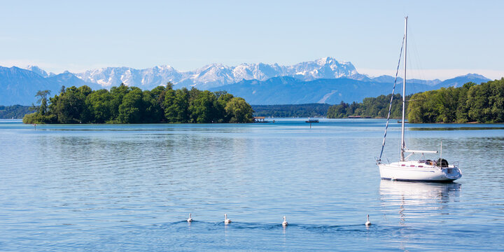 Scenic landscape of Lake Starnberg with Roseninsel (rose island) and an anchoring sailboat. Snow capped mountains at the horizon.