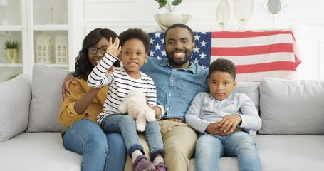 Portrait of young happy African American parents children sitting on sofa with USA flag and smiling cheerfully to camera. Joyful kids with mother and father on couch in living room. - Powered by Adobe