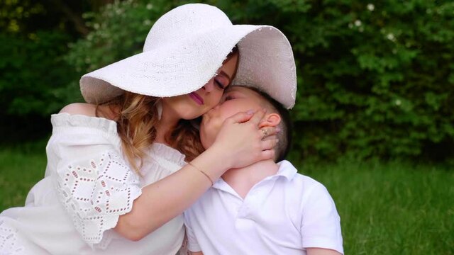 Mom and son hug, son kisses mom on the cheek, smiling. Woman in a white hat and little boy in a shirt are sitting in botanical garden on background of trees. 4K slow motion footage