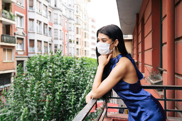 Beautiful model with mask looking at the street from her balcony in a blue dress