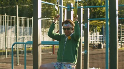 Funny freak man in green hoodie, color shorts and sunglasses pulling up on the rings on the sportground outdoors. Sport humor concept.