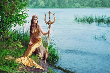 Fantasy woman real mermaid with trident myth goddess of sea with golden tail sitting in sunset on...