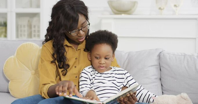 Young beautiful African American woman in glasses reading book with little cute daughter on sofa at home. Small child flipping pages of fairytale with pictures with mother on couch. Babysitter.