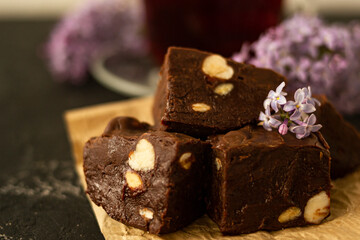 A piece of homemade chocolate cake on crumpled craft paper, with nuts on a black texture table. A cup of tea and lilac flowers decorate a delicious tea party.