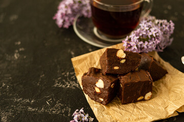 A piece of homemade chocolate cake on crumpled craft paper, with nuts on a black texture table. A cup of tea and lilac flowers decorate a delicious tea party.