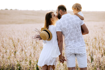 Happy family portrait outdoor at the sunset time. People having fun on the field. Concept of friendly family and of summer vacation. Parents and son spending good time. White dress. Straw hat. Kissing