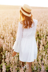 Back view of a pretty young woman in white dress holding straw hat running walking at the sage flower field. Beautiful girl enjoying a field of flowers, relaxing outdoors, having fun, harmony concept.