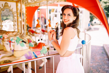 Fototapeta na wymiar Beautiful brunette girl in and the amusement park on the background of carousels with candy caramel in retro pin-up style
