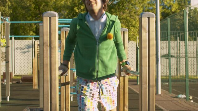 Funny freak man in green hoodie, color shorts and sunglasses pushing up on parallel bars on the sportground outdoors. Sport humor concept.